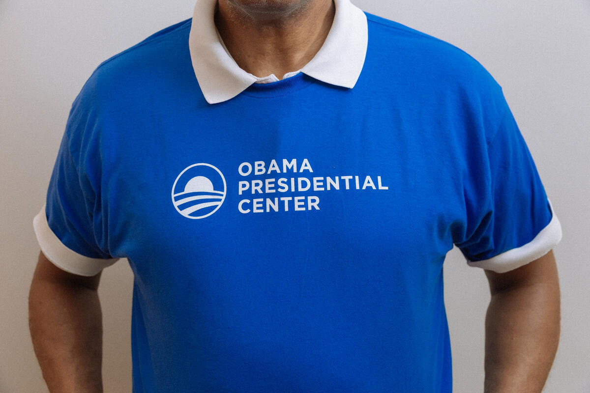 A person wears the Obama Presidential Center Logo T-shirt in blue with a white collar and cuffs, featuring the logo and text on the front. Their face isn’t visible. This shirt represents the Obama Foundation’s Chicago South Side center.