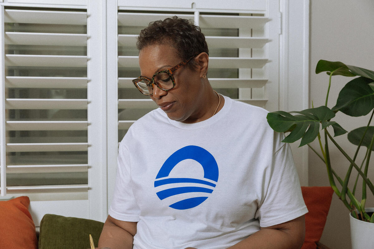 A woman wearing glasses and an Obama Presidential Center Symbol T-shirt with a bold blue graphic sits indoors by a window with white blinds and a green plant, looking down as she reads or writes.