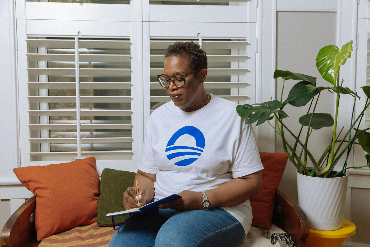 A person in glasses and an Obama Presidential Center Symbol T-Shirt – White sits on a cushioned bench, writing in a notebook. Nearby are pillows, a potted plant, and windows with white shutters behind them.