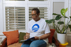 A person wearing glasses and the Obama Presidential Center Symbol T-shirt in white, featuring a blue logo, sits on a cushioned bench while writing in a notebook. A potted plant and white shutters are visible in the background.