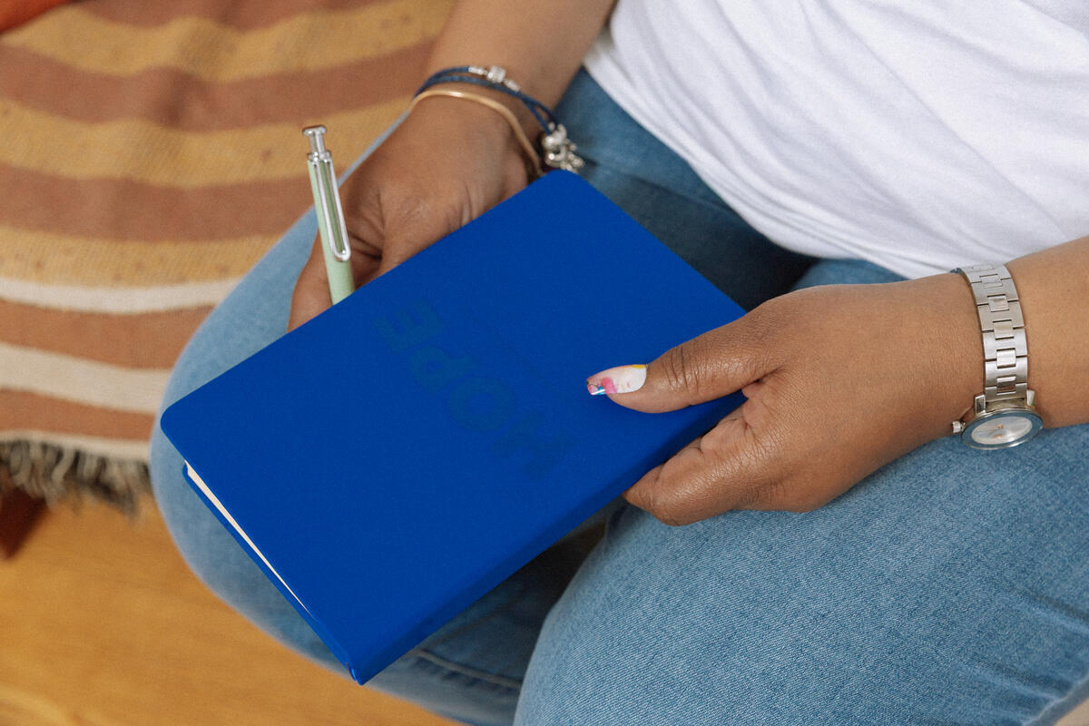 Wearing jeans and a watch, a person with stylish white and red nails holds the Obama Presidential Center Hope Journal in navy blue, featuring an embossed HOPE wordmark, ready to write.