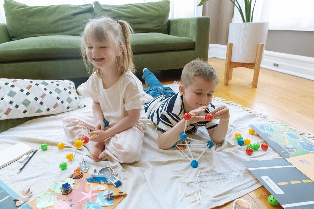 Two young children build structures with the Architecture and Engineering STEAM Kit from a Black-owned toy company, playing on a blanket indoors. The girl smiles as the boy concentrates, with a green couch and potted plant in the background.