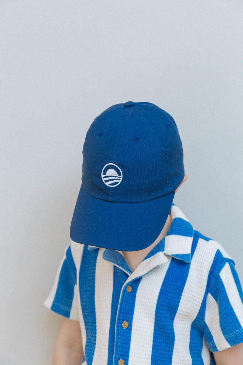 A young child in a blue and white striped shirt wears the Obama Presidential Center Symbol Cap, featuring a white circular logo, standing against a plain light background with the cap’s brim mostly covering their face.