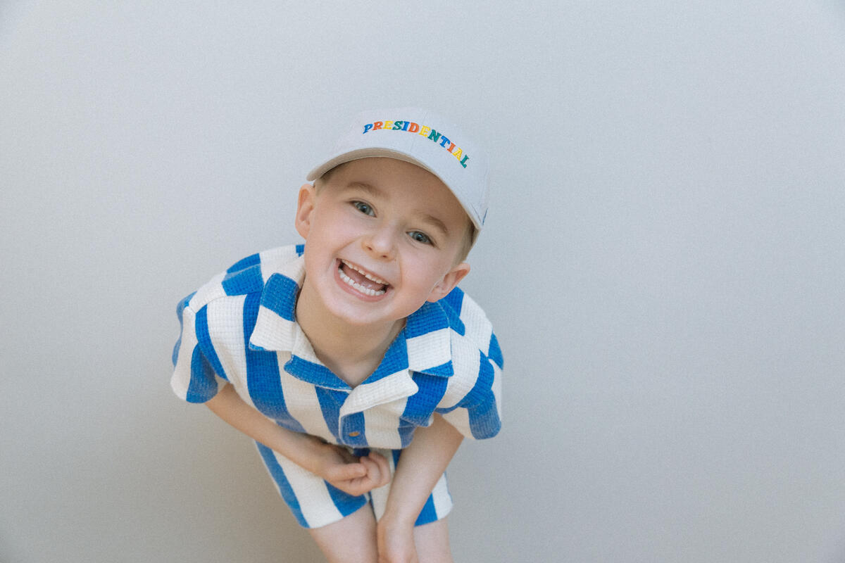 A smiling child wears the Presidential Embroidered Youth Cap in light gray with colorful PRESIDENTIAL letters, paired with a blue-and-white checkered outfit—an ideal look for the future Obama Foundation leader.