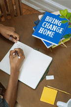 At a wooden table, someone writes in an Obama Presidential Center Bring Change Home Notebook, with blue versions of the same notebook, a plant, sticky notes, and a pencil nearby.