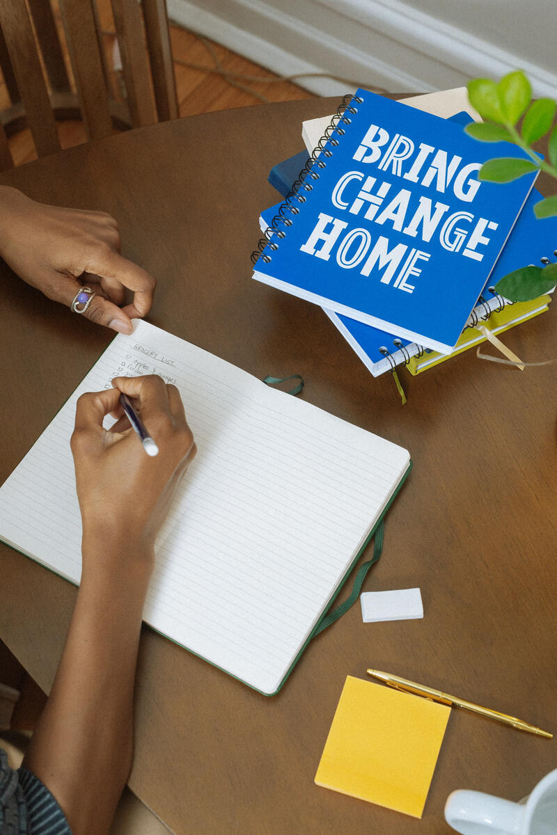 At a wooden table, someone writes in an Obama Presidential Center Bring Change Home Notebook, with blue versions of the same notebook, a plant, sticky notes, and a pencil nearby.