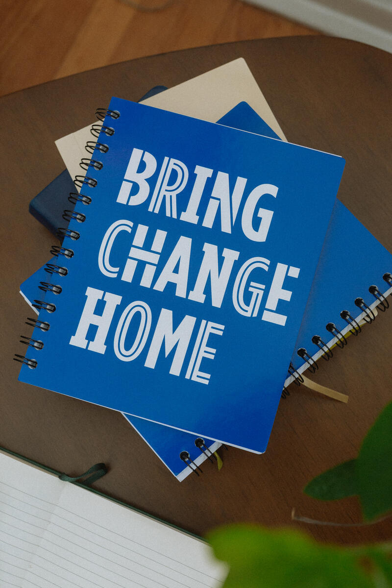 An Obama Presidential Center Bring Change Home Notebook with bold white lettering is stacked on two other notebooks on a wooden surface, with part of an open notebook and a green plant also visible.
