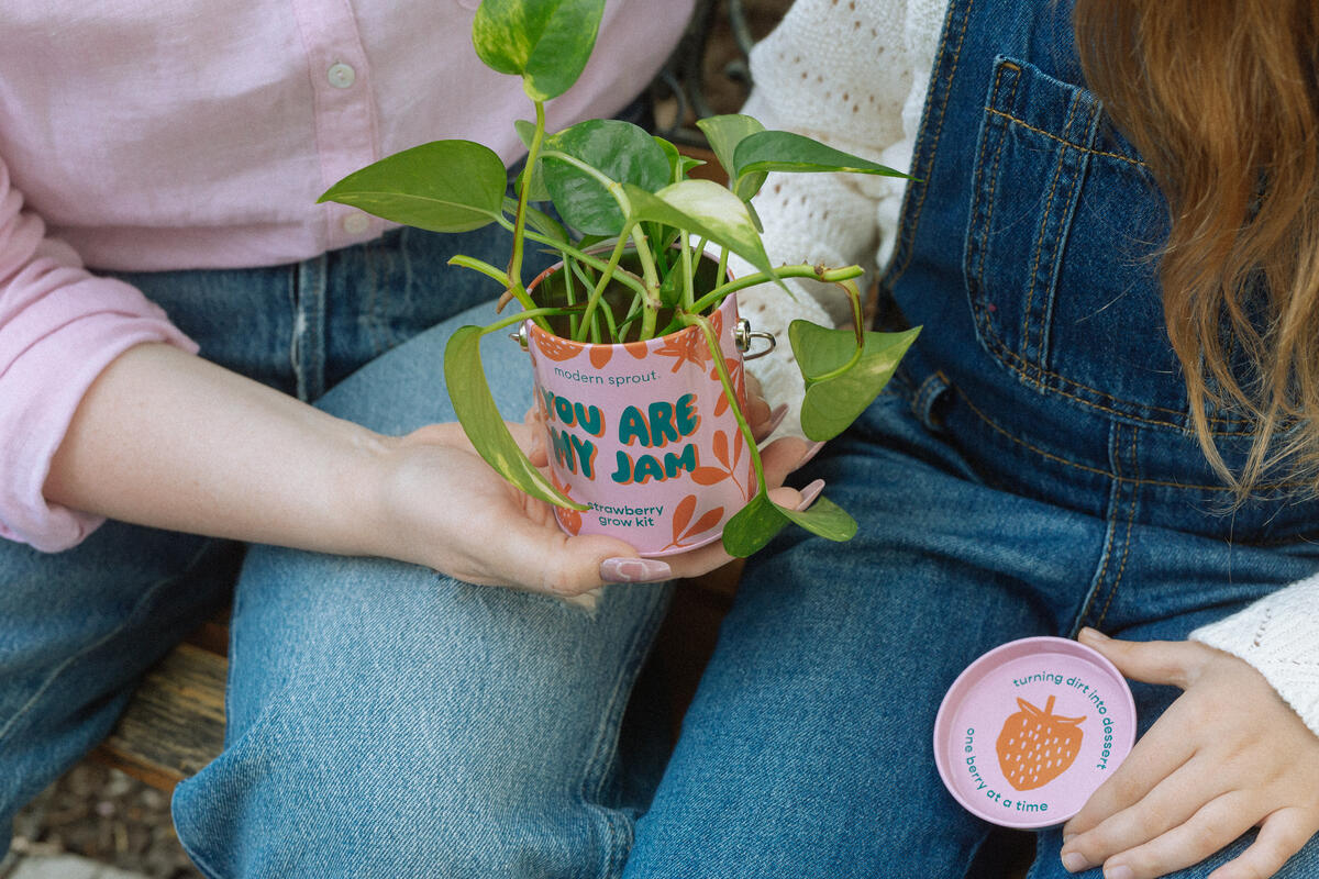 Two people sit side by side, each holding part of the "You Are My Jam Grow Kit"—one with the pink reusable planter and a small plant, the other with its matching lid. Only their jeans-clad torsos and hands are visible.