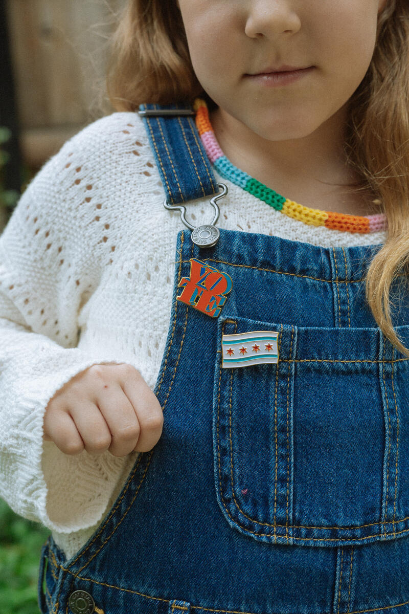 A young child wears denim overalls with two pins—one shaped like the Chicago flag and another resembling the Vote Pin—over a white knit sweater and a colorful beaded necklace, holding an overall strap.