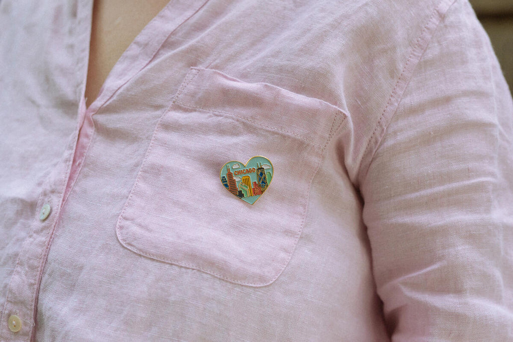 A person wears a light pink button-up shirt with the Skyline Heart Pin—a colorful enamel heart shaped like Chicago’s skyline and featuring the words SHOW UP—attached to the chest pocket.