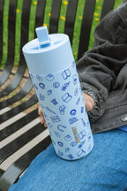 On Chicago’s South Side, a person in a dark jacket and blue jeans sits on a black metal bench with grass and yellow flowers behind them, holding an Obama Presidential Center Hope Water Bottle decorated with blue doodles.