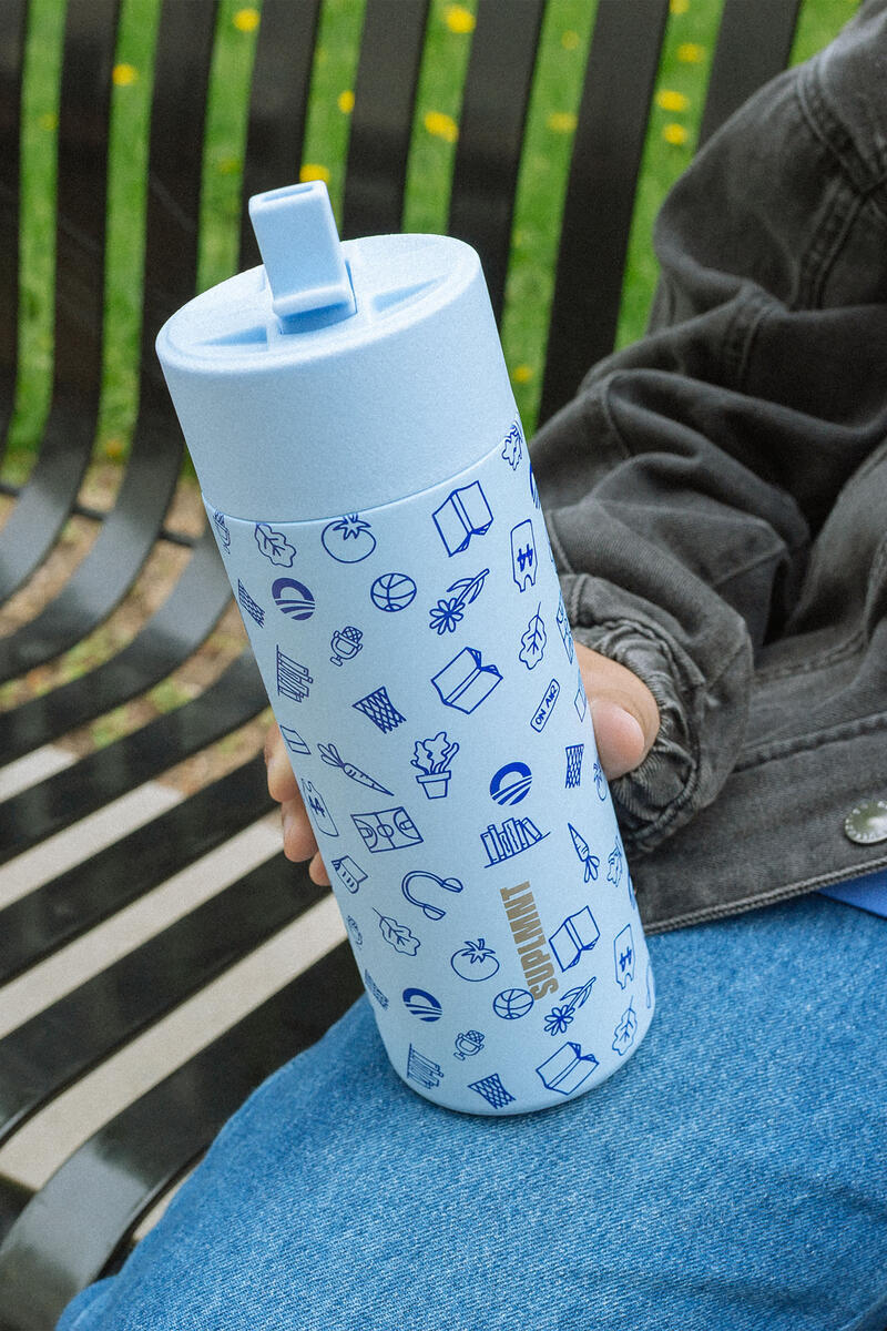 On Chicago’s South Side, a person in a dark jacket and blue jeans sits on a black metal bench with grass and yellow flowers behind them, holding an Obama Presidential Center Hope Water Bottle decorated with blue doodles.