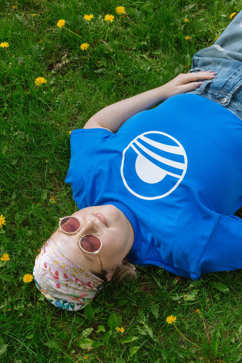 Wearing an Obama Presidential Center Symbol T-shirt, sunglasses, and a vibrant headscarf, a person relaxes on dandelion-speckled grass—capturing the ‘Bring Change Home’ spirit of the Obama Foundation.