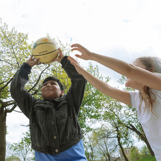 A boy holds the Obama Presidential Center Floral Basketball above his head as a girl tries to block him, playing outside amid trees and a cloudy sky, reflecting the vibrant energy of the Home Court collection.