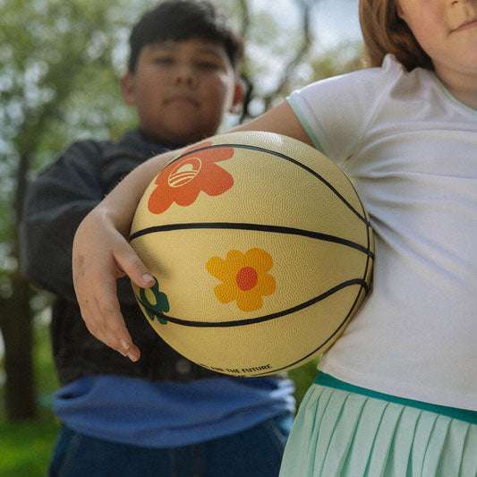A close-up captures two children outdoors, one holding the Obama Presidential Center Floral Basketball. The background is blurred with greenery, and only parts of their faces and bodies are visible, highlighting a spontaneous collaborative moment.