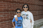 Two smiling children wearing sunglasses stand arm in arm in front of a brick wall. One wears a blue Obama Presidential Center Stickers Youth T-Shirt; the other wears a white hoodie that says It’s Up To Us.