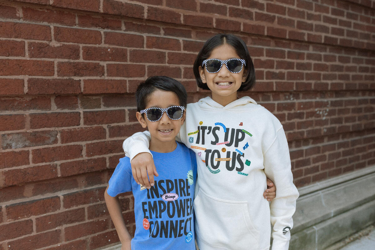 Two smiling children wearing sunglasses stand arm in arm in front of a brick wall. One wears a blue Obama Presidential Center Stickers Youth T-Shirt; the other wears a white hoodie that says It’s Up To Us.