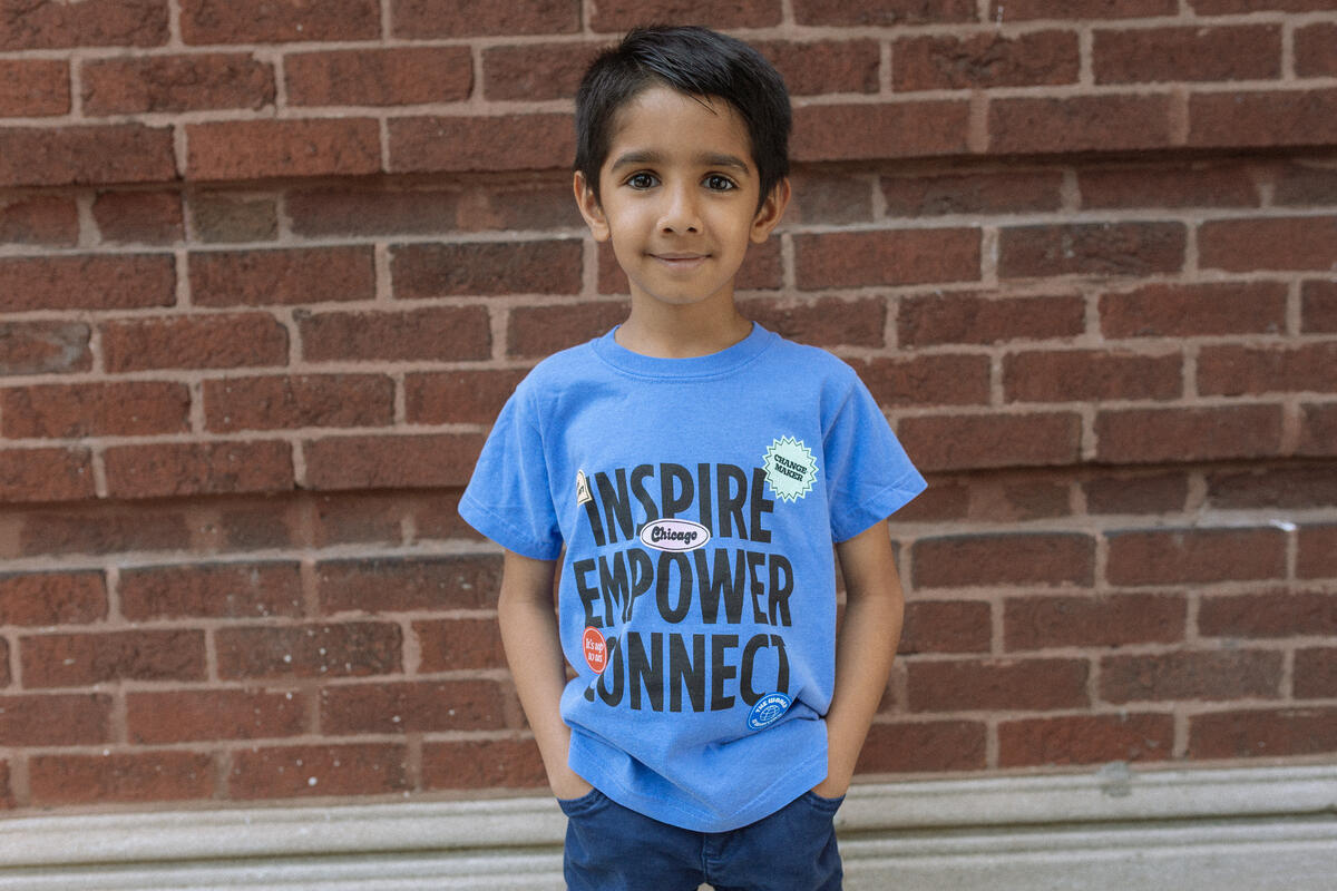 A young child stands against a brick wall, hands in pockets, wearing the Obama Presidential Center Stickers Youth T-Shirt that reads “Inspire Empower Connect,” adorned with various pins.