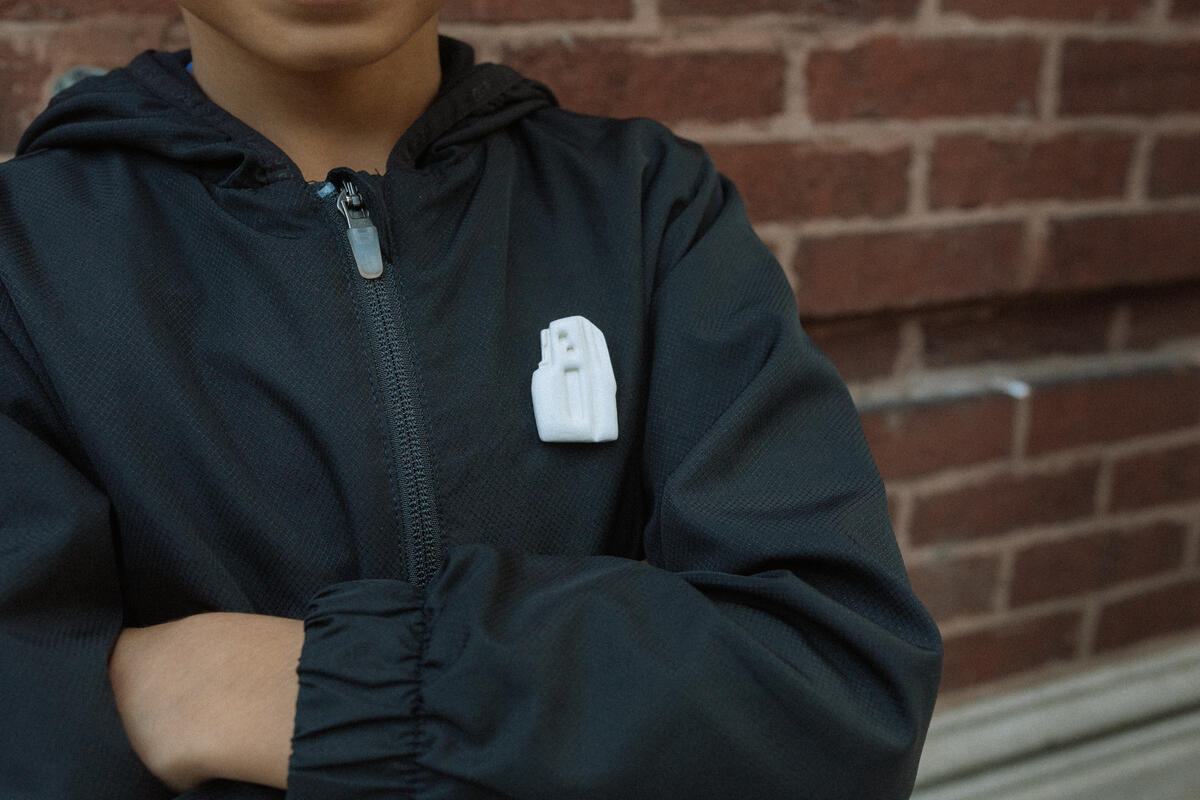 A child in a dark jacket with crossed arms stands before a brick wall. The jacket’s chest is adorned with an Obama Presidential Center Building Lapel Pin—small and white in color.