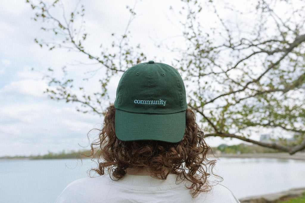 Someone with curly hair stands by a lake, facing away, wearing the Obama Presidential Center Community Cap with tree branches and water in the background.