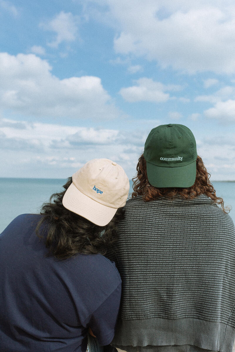Two people stand by the water, backs to the camera. One wears the beige Obama Presidential Center Hope Cap; the other a green cap labeled "community." Partly cloudy skies set a serene scene reflecting Obama Foundation values.