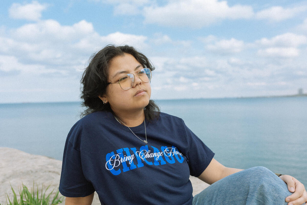 A person with glasses and long hair sits on a stone surface by a calm body of water, wearing the Obama Presidential Center Chicago Script T-Shirt in navy blue. The sky is partly cloudy, with a distant cityscape barely visible—possibly near the future site of the Obama Presidential Center.