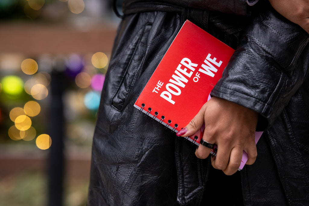 A person in a black leather jacket holds an Obama Foundation Notebook beside their leg, with blurry lights and decorations in the background.