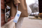 A person in a tan coat and white pants leans against a brick wall, holding an Obama Foundation Water Bottle with a blue logo and dotted pattern by its strap. Only part of another person and parked cars are visible in the background.