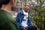 A young person in a denim jacket sits on outdoor steps, wearing the Obama Foundation Youth T-Shirt with "Bring Change Home" text. Green plants and a metal railing frame the scene.