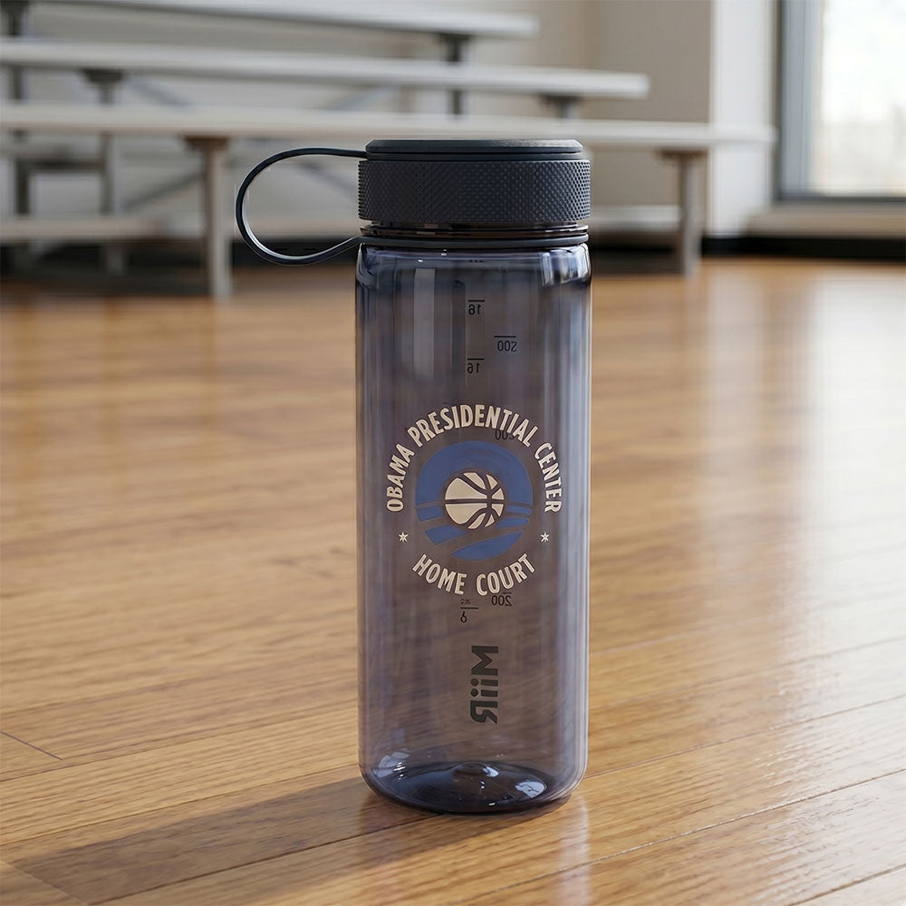 The Obama Presidential Center Home Court Water Bottle, a translucent design with a black lid and basketball graphic, sits on a wooden gym floor. Bleachers can be seen in the background. This is official Obama Foundation merchandise.