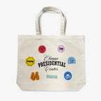The Obama Presidential Center Stickers Tote is a cream canvas bag featuring black "Obama Presidential Center" text and colorful patches like HOPE, 44, YES, It’s Up To Us!, Driving Change Home, and the Obama logo—official foundation merchandise.