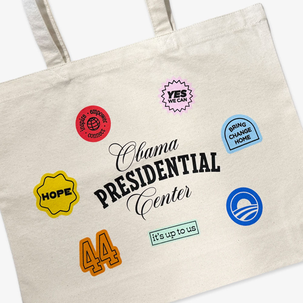 The Obama Presidential Center Stickers Tote is a cream canvas bag featuring black "Obama Presidential Center" text and colorful patches like HOPE, 44, YES, It’s Up To Us!, Driving Change Home, and the Obama logo—official foundation merchandise.