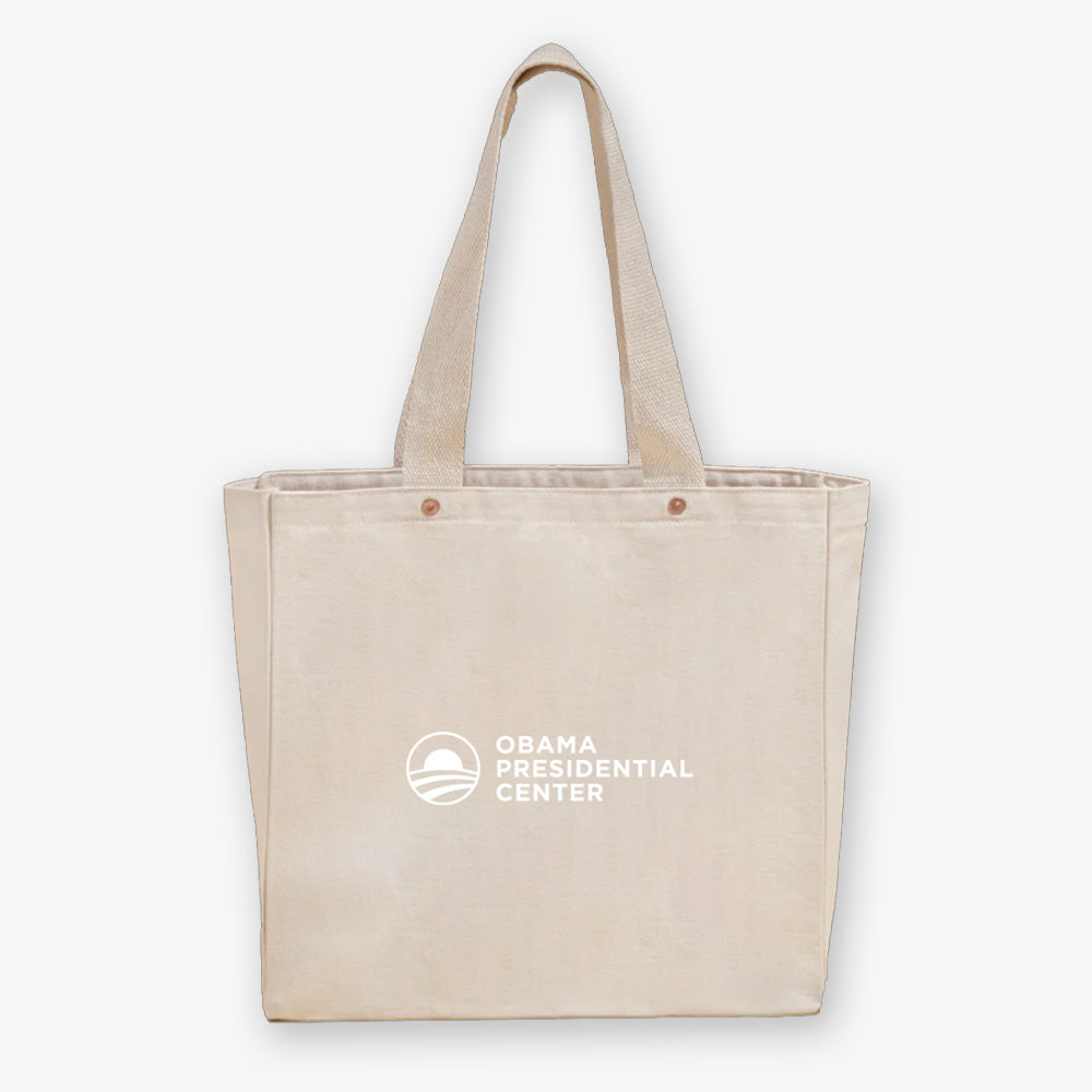 The Obama Presidential Center "You Are America" Speech Tote is a beige canvas bag with two handles, featuring the center’s logo and text in white on the front.