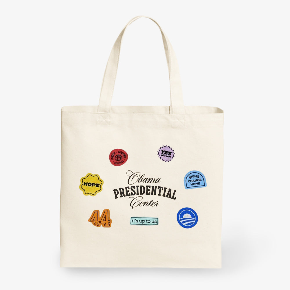 The Obama Presidential Center Stickers Tote is a cream canvas bag featuring black "Obama Presidential Center" text and colorful patches like HOPE, 44, YES, It’s Up To Us!, Driving Change Home, and the Obama logo—official foundation merchandise.