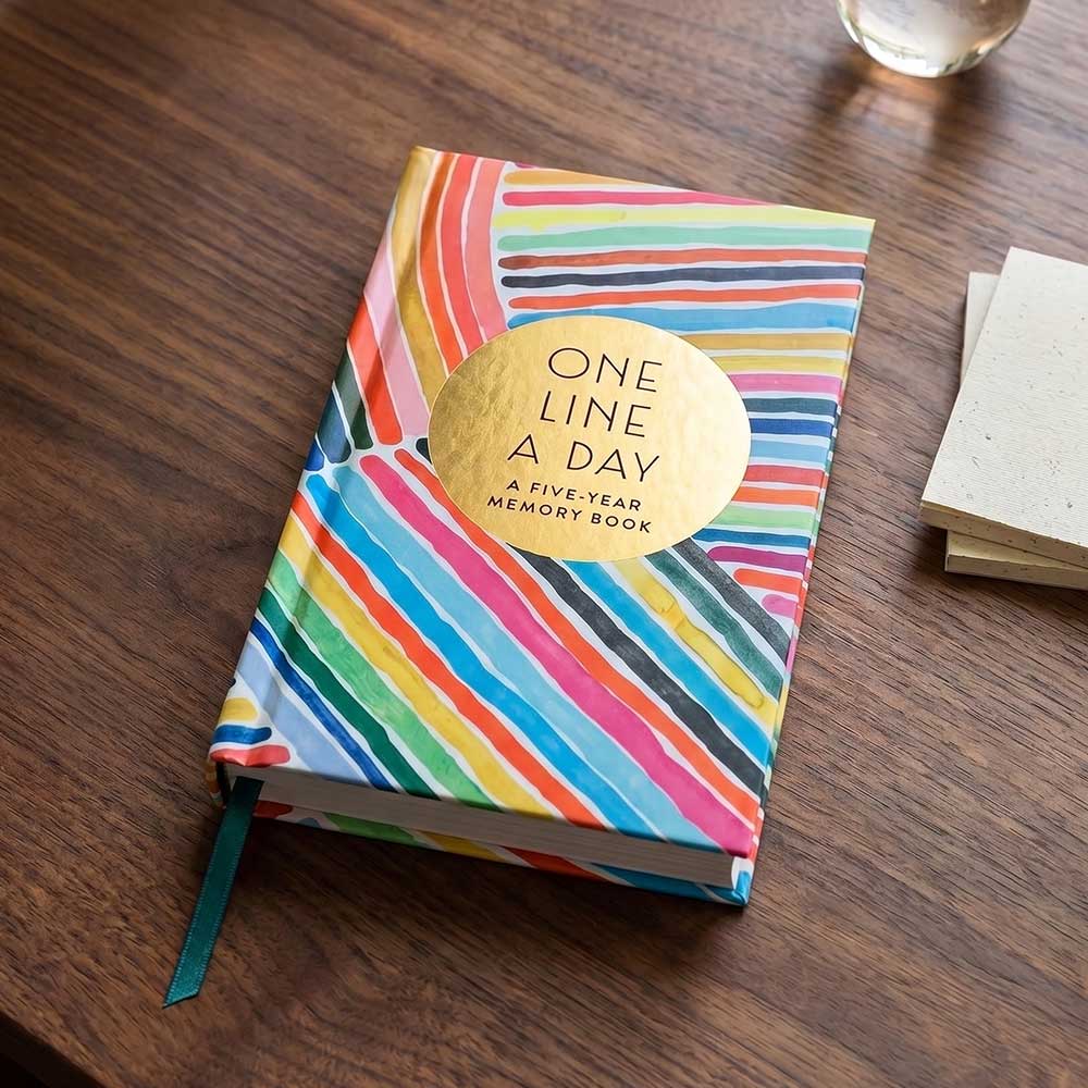 The Rainbow One Line a Day: A Five-Year Memory Book, featuring rainbow stripes and a gold circle on the cover, rests on a table next to a drink and notepads—perfect for capturing daily memories over five years.
