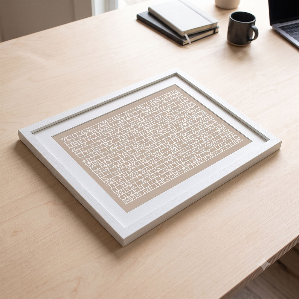 A framed Obama Presidential Center "You Are America" Speech Matted Print with abstract geometric white lines rests on a light wooden desk beside a closed notebook, laptop, and dark coffee mug. Sunlight streaming through a window in the background fills the workspace with inspiration, echoing the creative spirit of the Obama Foundation.