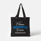 The Obama Presidential Center Script Tote is a black canvas bag with "Obama Presidential Center" in white and blue text, plus "Inspire Empower & Connect" and Obama Foundation locations printed in small white font at the bottom.