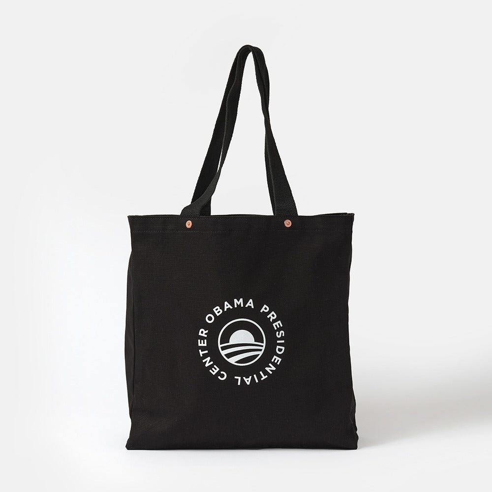 The Obama Presidential Center Script Tote is a black canvas bag with two handles, featuring a white circular logo with "Obama Presidential Center" around a stylized O, shown against a plain white background.
