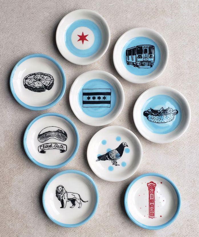 Nine small handmade porcelain Pigeon Trinket Dishes are arranged on a beige surface, each featuring blue, red, or black illustrations of iconic Chicago symbols—including deep dish pizza, a train, flags, local foods, a pigeon motif, the Art Institute lion, “Cloud Gate,” and a street sign—making them the perfect Chicago gift.
