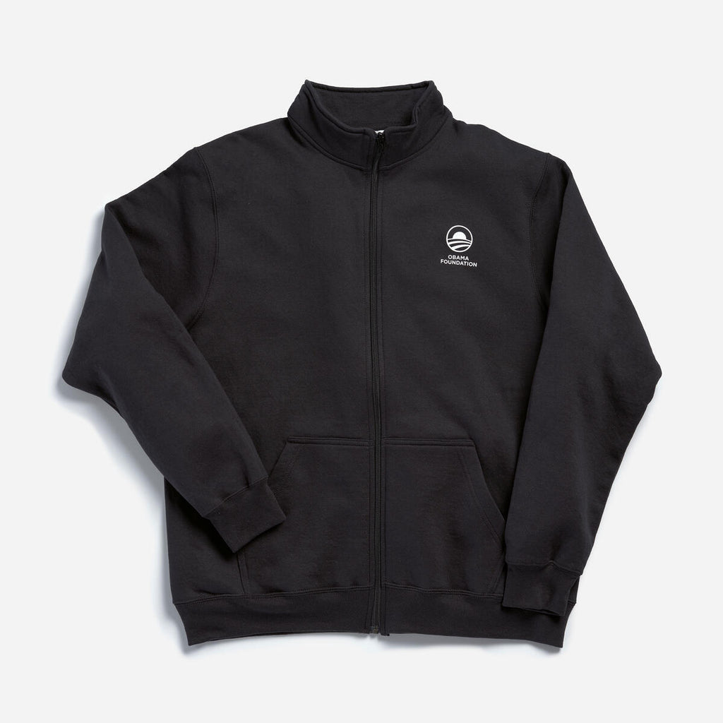 The Obama Foundation Full-zip Fleece is a black fleece jacket with a high collar, small white logo, and "Obama Foundation" text on the left chest, full zip closure, long sleeves, and two front pockets.