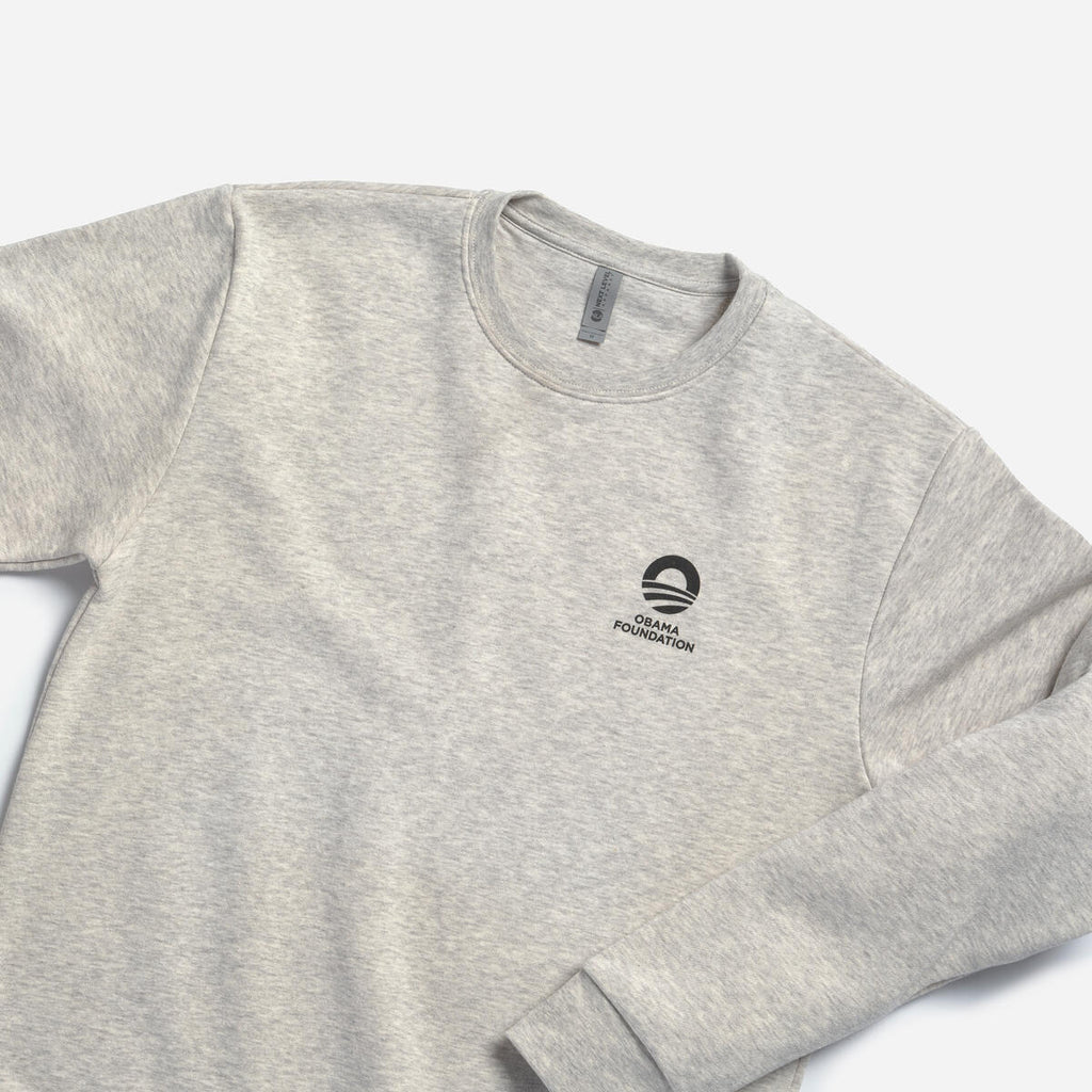 The Obama Foundation Sweatshirt is an oatmeal heather crewneck with the foundation's logo and text in black on the left chest, shown against a white background.