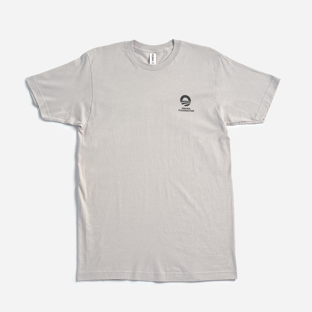 The Obama Foundation T-Shirt is a plain light gray crewneck laying flat on a white surface, featuring a small black graphic and the Obama Foundation’s rising sun logo with text on the left chest.