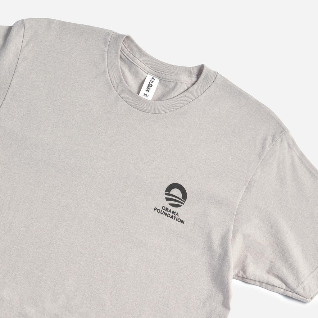The Obama Foundation T-Shirt is a light gray crewneck tee with the rising sun logo and black text printed on the left chest, shown laid flat on a white background.