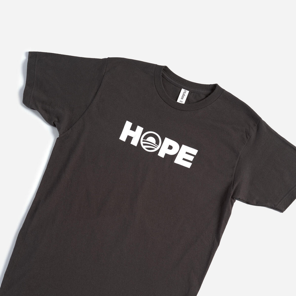 The Obama Foundation "Hope" T-shirt is black with bold white HOPE lettering; the O has a rising sun logo with red and blue stripes, inspired by the classic Obama Foundation design.