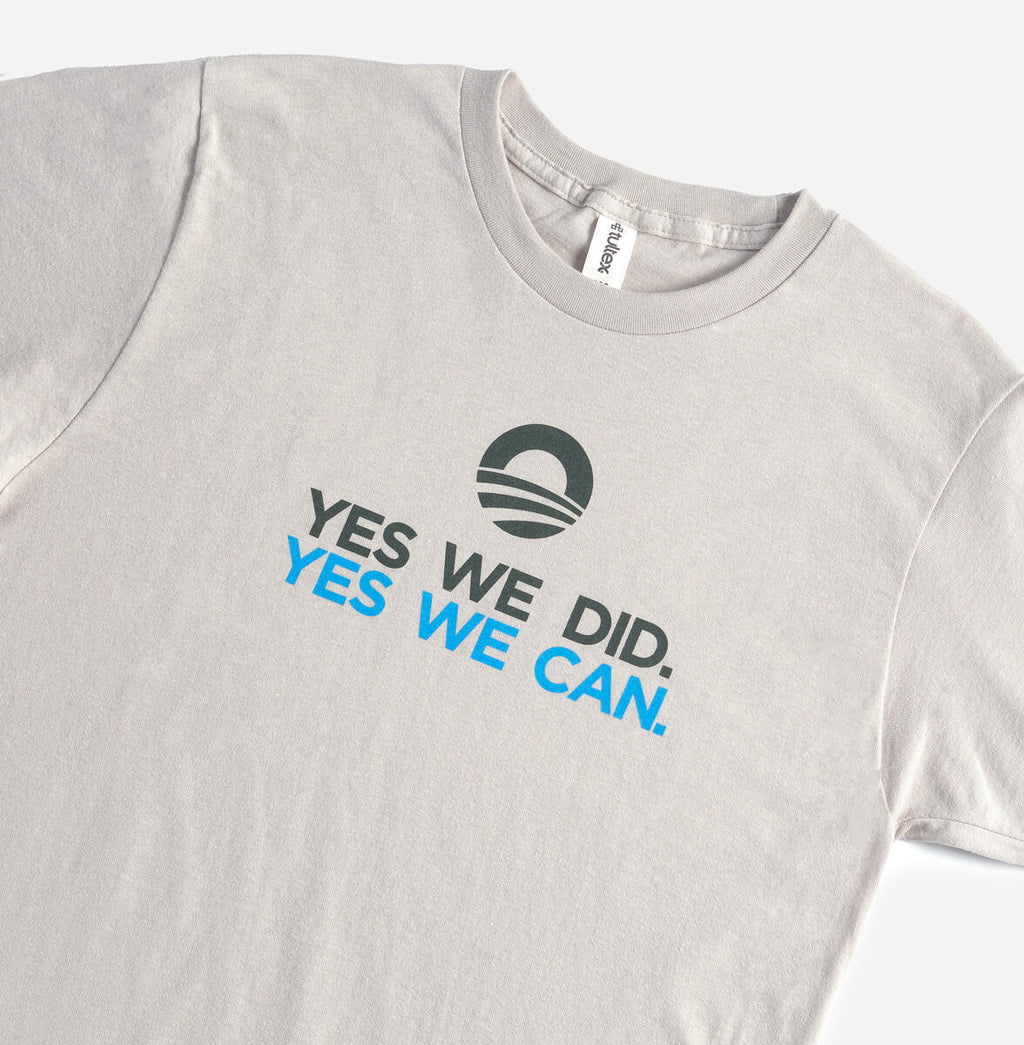 The "Yes We Can" T-shirt is a light gray shirt featuring the Obama Foundation campaign logo above “YES WE DID.” in black and “YES WE CAN.” in blue, celebrating Barack Obama's iconic farewell address.