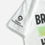 Close-up of the Obama Foundation Youth T-Shirt sleeve, featuring the black Obama Foundation logo and name. The front displays bold black text and a green-and-yellow Bring Change Home graphic.